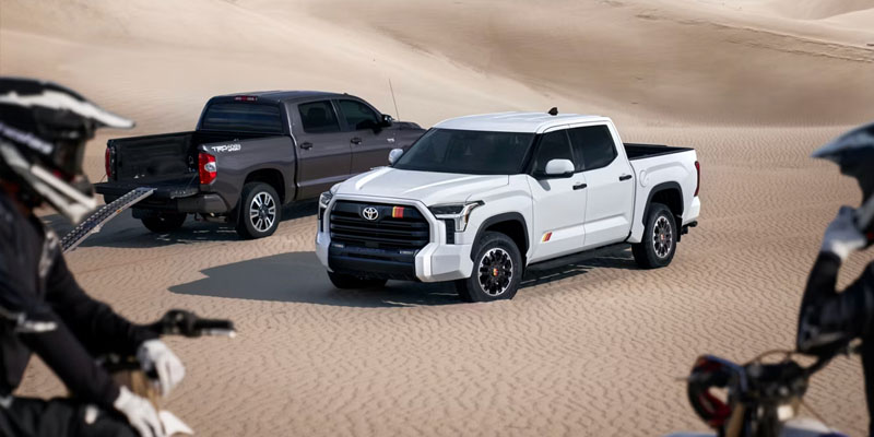 Two Toyota pickup trucks driving across sandy desert dunes, with off-road motorcycles in the foreground, highlighting rugged performance and adventure capability.